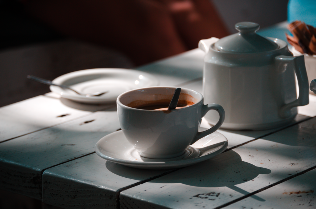 A white ceramic coffee cup and pot on a sunlit painted wooden table.
